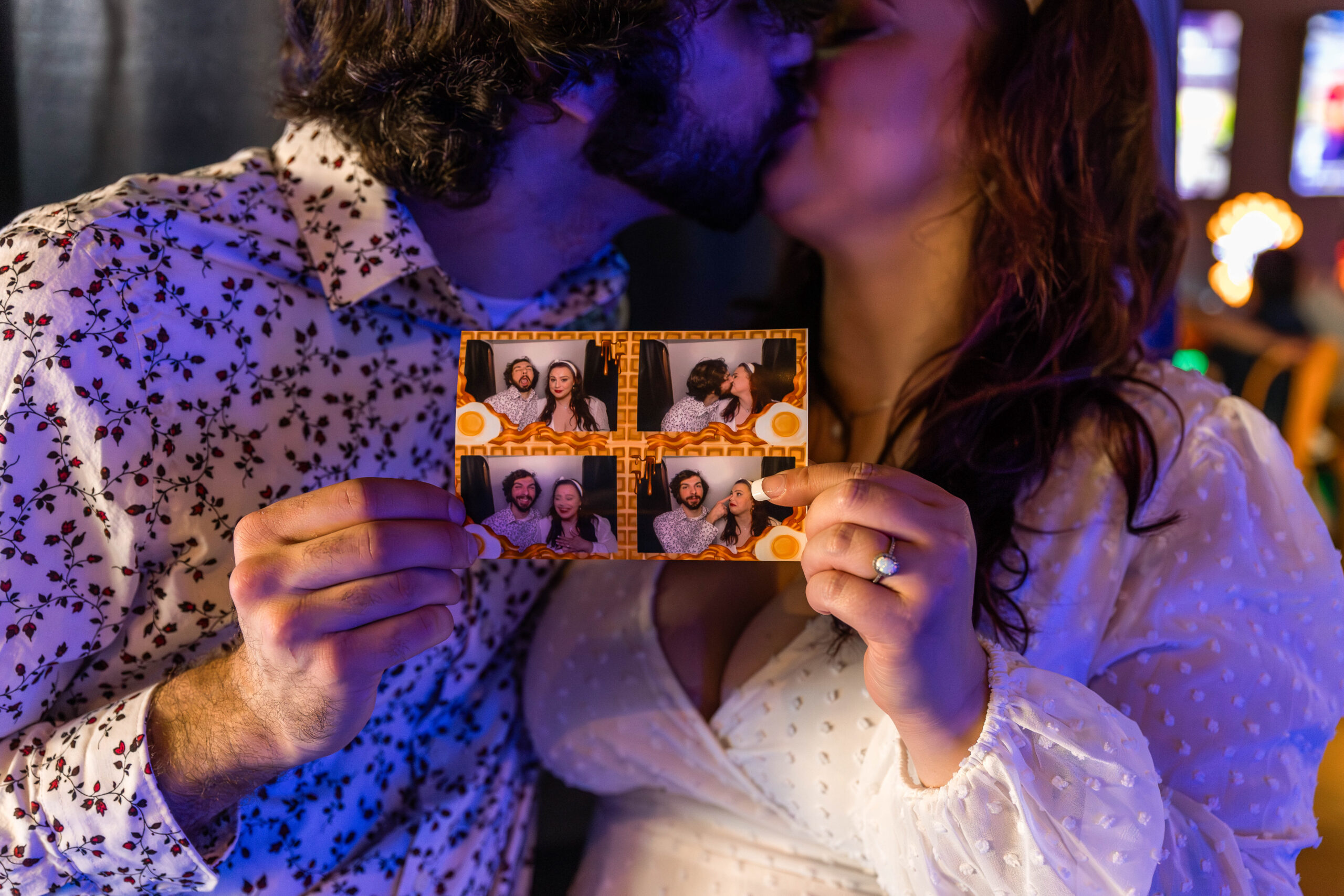 A couple kisses while holding up a photo booth strip featuring four playful snapshots of them together. The image is set in a fun, arcade-style environment with vibrant lights in the background. The woman's engagement ring is visible as she holds the photo strip, adding a personal and romantic touch to the moment.