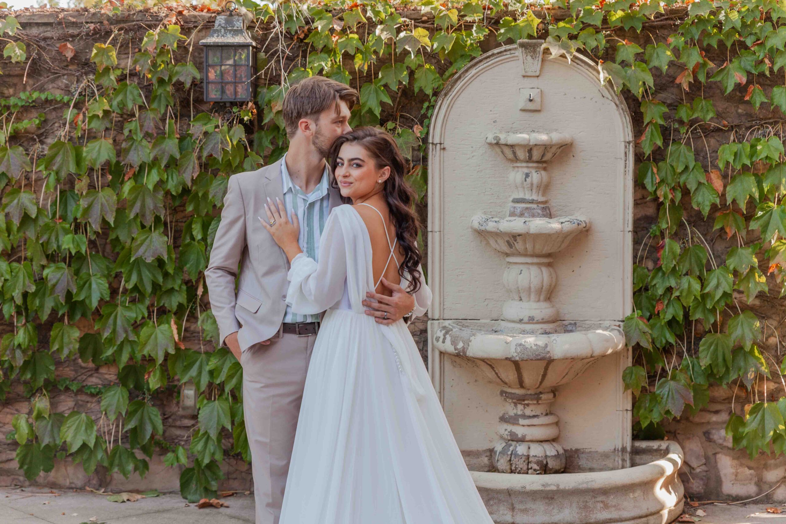 Bride and groom embracing beside a vintage stone fountain with ivy-covered wall in the background.