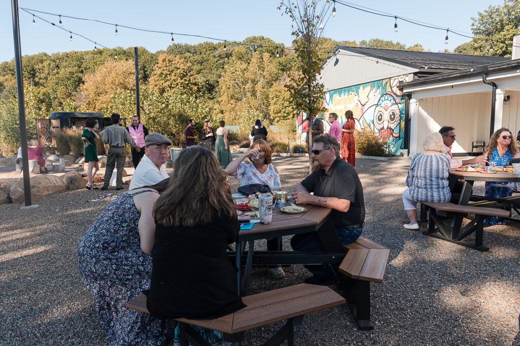 Wedding guests enjoying cocktail hour at Himelright Lodge with picnic tables, string lights, and fall trees at Cascade Valley Metro Park.